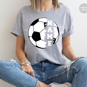 May include: Heather grey t-shirt with a black and white soccer ball graphic. The soccer ball is split in half, with the word "NAME" written vertically down the side of the ball.