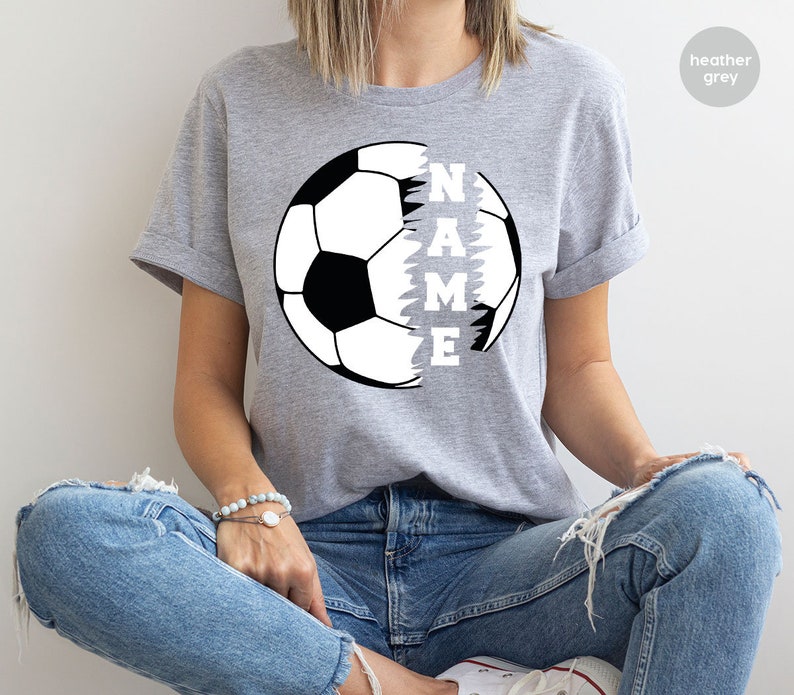 May include: Heather grey t-shirt with a black and white soccer ball graphic. The soccer ball is split in half, with the word "NAME" written vertically down the side of the ball.