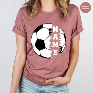 May include: Heather mauve t-shirt with a white soccer ball graphic and the word "NAME" written vertically down the side of the ball.