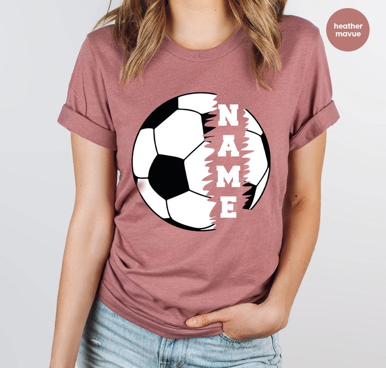 May include: Heather mauve t-shirt with a white soccer ball graphic and the word "NAME" written vertically down the side of the ball.