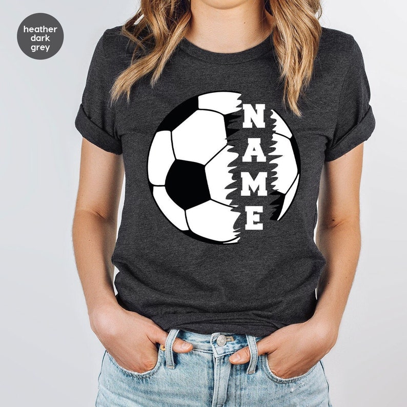 May include: Heather dark grey t-shirt with a white soccer ball graphic. The word "NAME" is written vertically inside the ball, with each letter appearing in a different section of the ball.