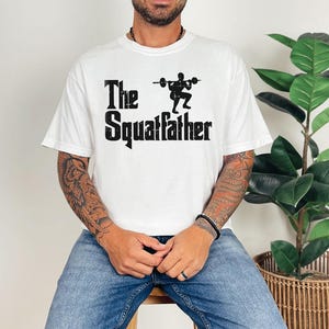 Peut inclure: T-shirt blanc avec un texte noir qui dit "The Squatfather" et une silhouette d'une personne soulevant des poids.