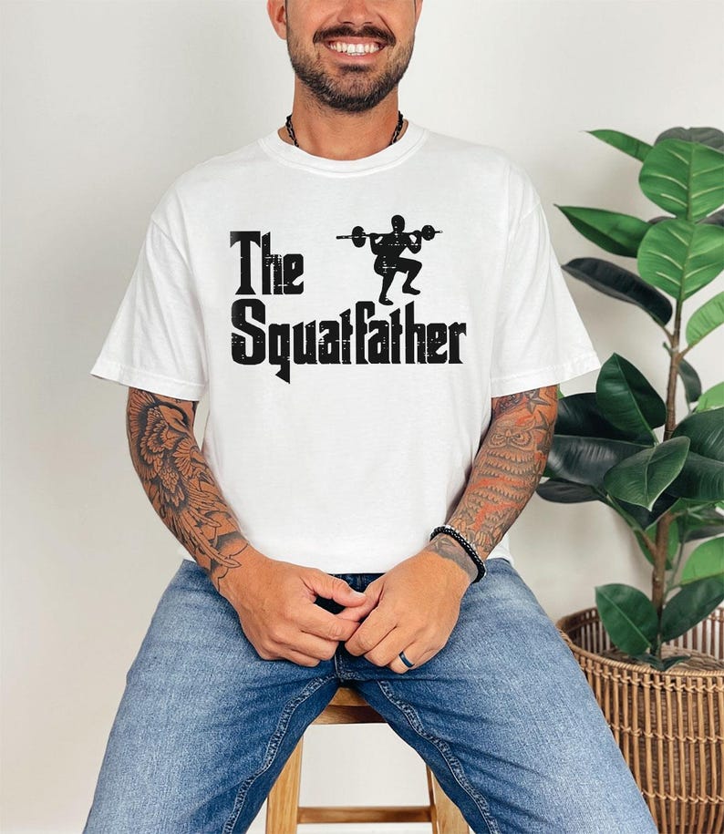 Peut inclure: T-shirt blanc avec un texte noir qui dit "The Squatfather" et une silhouette d'une personne soulevant des poids.
