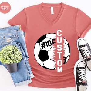 May include: Heather mauve V-neck t-shirt featuring a soccer ball graphic with the word "CUSTOM" written vertically down the side of the ball. The soccer ball has the number "#10" on it.