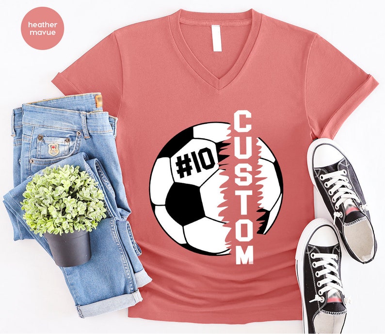 May include: Heather mauve V-neck t-shirt featuring a soccer ball graphic with the word "CUSTOM" written vertically down the side of the ball. The soccer ball has the number "#10" on it.