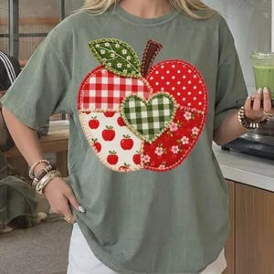 May include: A sage green short-sleeve t-shirt featuring a large central graphic of an apple made from various fabric-style patterns. The apple design includes red and white gingham, red with white polka dots, small red apples, and floral prints.