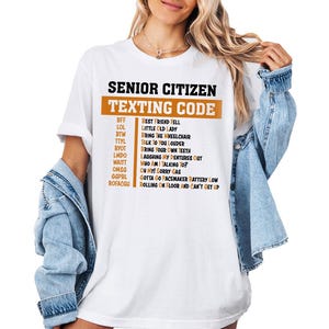 May include: White t-shirt with a black and orange graphic that says "Senior Citizen Texting Code" and lists common senior citizen texting abbreviations and their meanings.