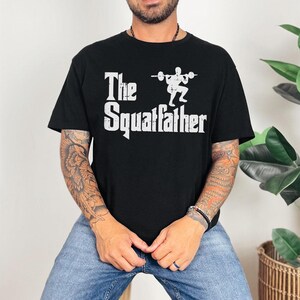 Peut inclure: Un t-shirt noir avec un imprim&eacute; graphique blanc qui dit "The Squafather" avec une silhouette d'une personne soulevant des poids.
