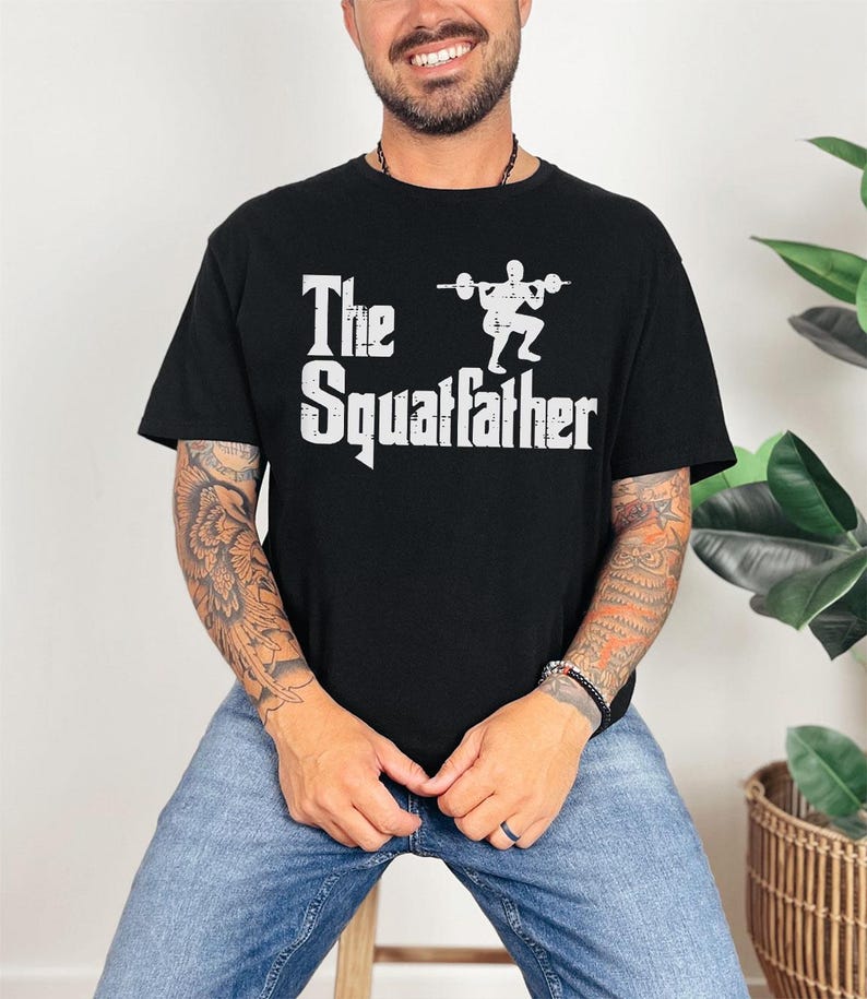 Peut inclure: Un t-shirt noir avec un imprim&eacute; graphique blanc qui dit "The Squafather" avec une silhouette d'une personne soulevant des poids.
