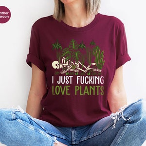 May include: A heather maroon t-shirt with a graphic of a skeleton lying in a bed of green plants. The text "I JUST FUCKING LOVE PLANTS" is printed in white below the graphic.