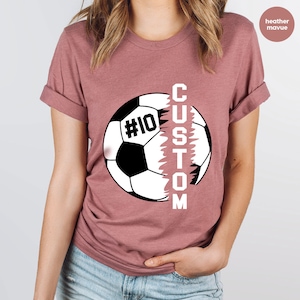 May include: Heather mauve t-shirt featuring a soccer ball graphic with the word "CUSTOM" printed vertically down the side of the ball. The soccer ball has the number "#10" printed on it.