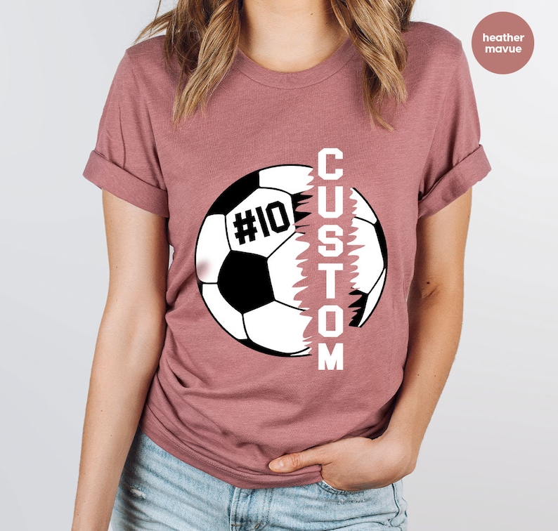 May include: Heather mauve t-shirt featuring a soccer ball graphic with the word "CUSTOM" printed vertically down the side of the ball. The soccer ball has the number "#10" printed on it.