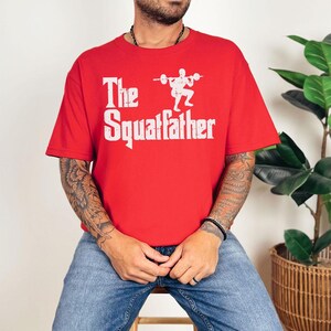 Peut inclure: Un t-shirt rouge avec un imprim&eacute; graphique blanc d'une personne accroupie avec une barre. Le texte "The Squatfather" est imprim&eacute; sous l'image.