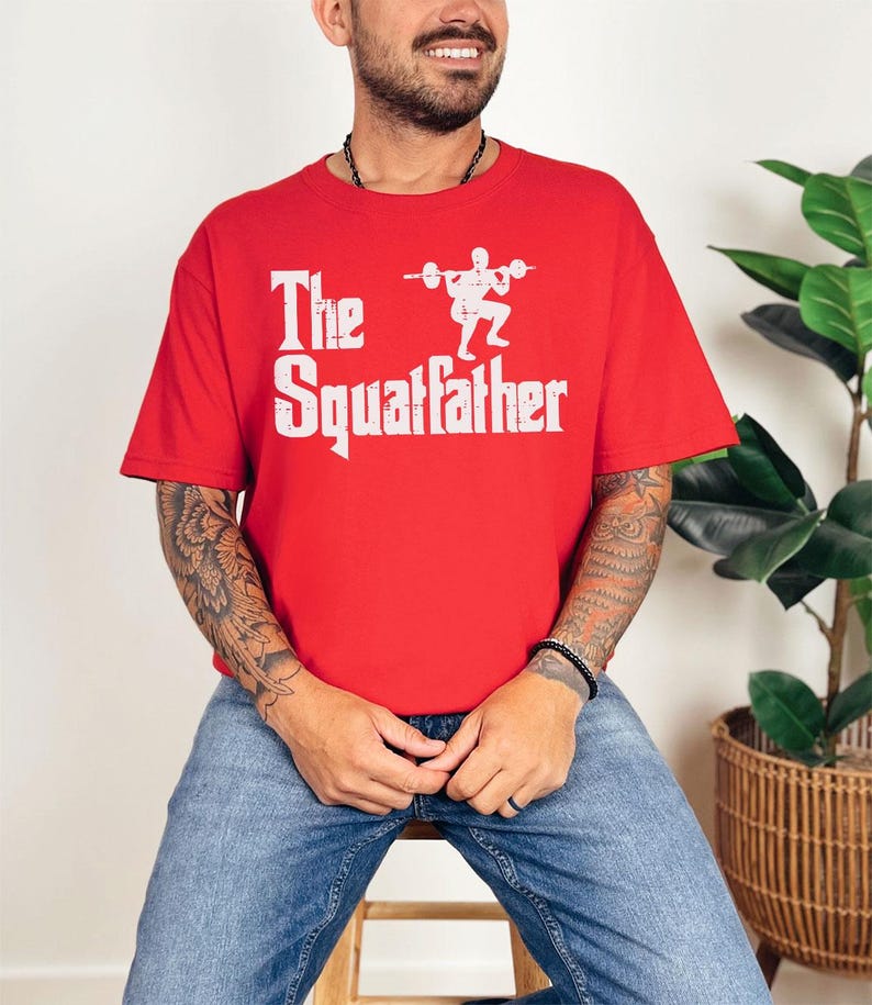 Peut inclure: Un t-shirt rouge avec un imprim&eacute; graphique blanc d'une personne accroupie avec une barre. Le texte "The Squatfather" est imprim&eacute; sous l'image.