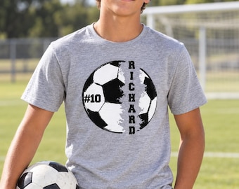 Può includere: Una t-shirt grigio melange a maniche corte con una grande grafica di un pallone da calcio bianco e nero. La grafica include il testo "RICHARD" in verticale e il numero "#10" stampato in nero sul lato sinistro del disegno del pallone.