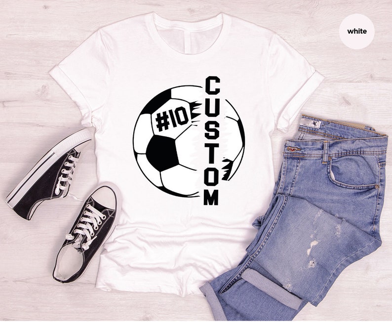 May include: White t-shirt featuring a black soccer ball graphic with the word "CUSTOM" written vertically down the side of the ball. The ball has the number "#10" on it.