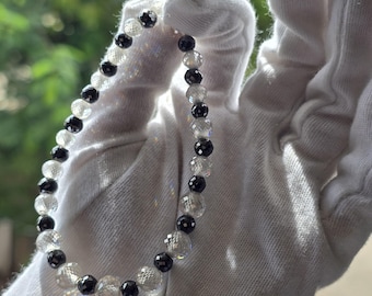 May include: A beaded bracelet featuring alternating clear and black faceted beads. The clear beads reflect light, creating a sparkling effect. The bracelet is held against a white background, highlighting the contrast between the beads.