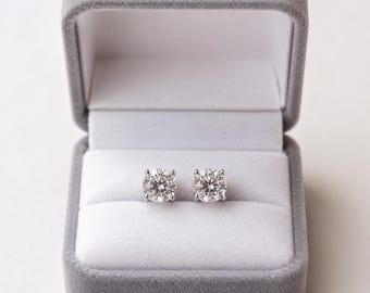 May include: A pair of round-cut stud earrings featuring clear, faceted stones set in silver-toned metal prongs. The earrings are displayed side-by-side within a grey velvet-lined jewelry box that has a hinged lid and a white interior cushion.