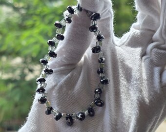 May include: A beaded necklace featuring black, faceted beads interspersed with smaller, clear beads. The necklace is held against a white background, with a blurred green backdrop.