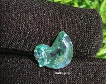 May include: A faceted, teal-colored gemstone with a unique, abstract shape. The gem is displayed on a dark, textured surface, with a blurred green background. The text "Rufkagems" is visible on the gem.
