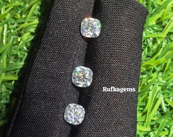 May include: Four cushion-cut gemstones displayed on a black fabric against a backdrop of green grass. The gemstones are clear and sparkling, with visible facets. The text "Rufkagems" is visible on the fabric.