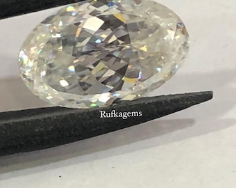 May include: An oval-cut, colorless gemstone with a brilliant faceted surface that reflects light, creating a sparkling effect. The stone is held by black tweezers, and the text Rufkagem is printed in small white letters on the lower edge.