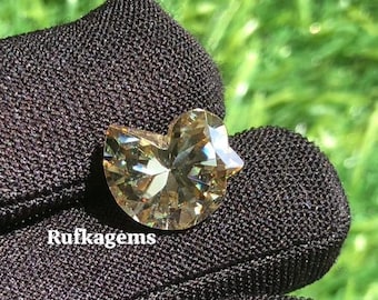 May include: A faceted, heart-shaped gemstone with a light brown hue. The gem is held against a dark, textured background, with the text "Rufkagems" visible. The background is blurred green.