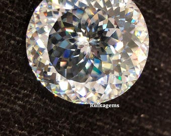 May include: A round, clear gemstone with a brilliant cut, reflecting a spectrum of colours. The facets of the stone are clearly defined. The image is set against a dark background, with the text "Rutkagems" visible.