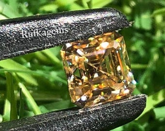 May include: A radiant, square-cut, golden-brown gemstone is held by a black tweezer against a backdrop of green foliage. The gemstone has multiple facets that reflect light. The text "Rufkagems" is visible on the tweezer.