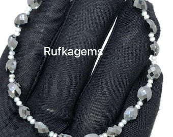 May include: A bracelet featuring faceted, dark gray gemstones and small white beads strung together. The bracelet is displayed on a black surface. The text "Rufkagems" is visible on the surface.