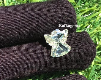 May include: A faceted, light blue gemstone angel figurine. The angel is resting on a dark brown fabric. The text "Rufkagems" is visible in the upper right corner. The background is green.