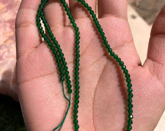 May include: A strand of small, faceted, emerald green beads. The beads are strung together on a green cord, forming a necklace or bracelet. The beads are held in a person's hand, showcasing their color and size.