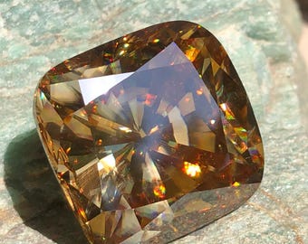 May include: A large, faceted, cushion-cut gemstone with a warm, golden-brown hue. The gem's surface reflects light, displaying a spectrum of colors. The stone is set against a textured, greenish-gray surface. The text "Rufkagems" is visible on the surface.