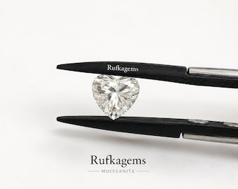 May include: A heart-shaped, clear, faceted moissanite gemstone held by black metal tweezers. The text "Rufkegems" is printed on the upper tweezer arm, and the text "Rufkegems MOISSANITE" appears below the gemstone against a plain white background.