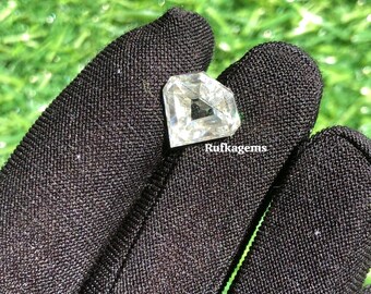 May include: A clear, faceted gemstone with a unique shape, held against a black fabric background. The gem has a central square cut-out and is slightly off-white in color. The text "Rufkagems" is visible.