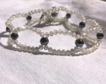 May include: A delicate, layered bracelet featuring clear, faceted beads and interspersed with dark, round beads. The bracelet is displayed on a white surface, showcasing its intricate design and sparkling appearance. The clasp is silver.