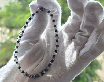 May include: A delicate bracelet composed of small, square beads in clear, black, and gray hues. The beads are strung together to form a flexible chain. The bracelet is held against a white background, showcasing its design.