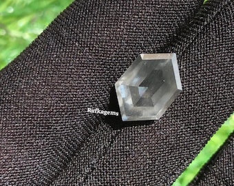 May include: A clear, hexagonal-shaped gemstone with multiple facets. The gem is held against a black fabric background, with the text "Rufkagems" visible. The background includes a blurred green area.
