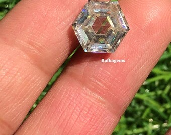 May include: A clear, hexagonal-cut gemstone with multiple facets, held between fingers. The gem has a complex internal structure, reflecting light and creating a rainbow effect. The word "Rufkagems" is visible.
