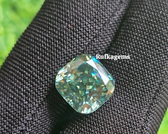 May include: A cushion-cut gemstone with a light teal hue, set against a black fabric background. The gem reflects light, displaying various shades of blue and green. The text "Rufkagems" is visible.