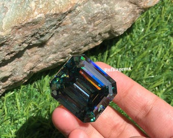 May include: A large, emerald-cut gemstone with a dark teal hue, displaying internal color flashes. The gem is held against a backdrop of green grass and a large rock. The text "Rufkagems" is visible on the side of the gem.