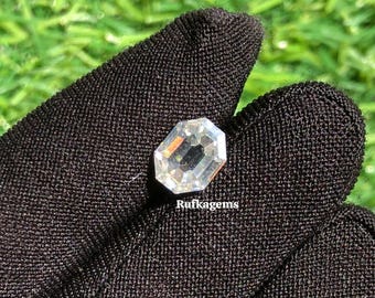 May include: A clear, faceted gemstone with an octagonal shape, held in a black glove. The gem reflects light, showcasing its internal structure. The background is blurred green grass. The text "Rufkagems" is visible.