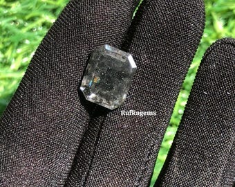 May include: A faceted, rectangular gemstone with a smoky, translucent appearance. The gem is held in a black glove, with the text "Rufkagems" visible. The background is blurred green.