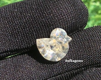 May include: A clear, faceted gemstone shaped like a duck. The gem is set against a black fabric background. The text "Rufkagems" is visible on the fabric. The duck-shaped gem is a unique and eye-catching piece.