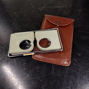 May include: A pair of magnifying glasses with a brown leather case. The magnifying glasses are silver and have a green plastic frame. The case has a flap with a button closure.