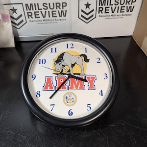 May include: A round black-framed clock with a white face featuring the word "ARMY" in red and blue. The clock face also displays a cartoon horse and the words "DEPARTMENT OF THE ARMY UNITED STATES OF AMERICA".