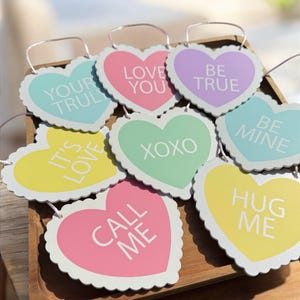 May include: A collection of heart-shaped decorations in various pastel colors, each with a different romantic message in white text. The hearts have scalloped edges and are displayed on a wooden tray.