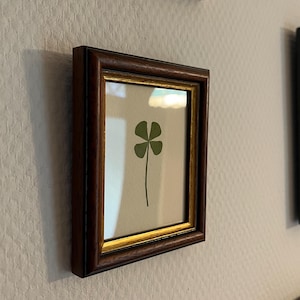 以下が含まれることがあります： 濃い茶色のフレームに金の縁取りが施された、四つ葉のクローバーのプリントが額装されています。