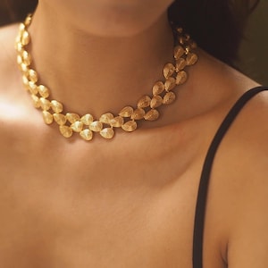 Może przedstawiać: Złoty naszyjnik z delikatnym wzorem nakładających się kształtów łez. Naszyjnik nosi osoba w czarnym topie na ramiączkach.