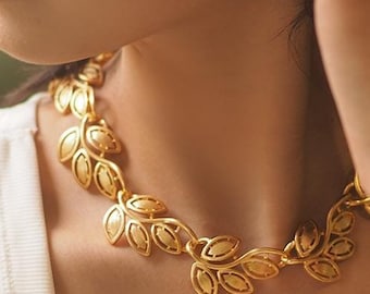 May include: A gold-colored necklace with a leaf design. The necklace is worn on a person wearing a white tank top. A silver bracelet is also visible on the person's wrist. The necklace is the focal point of the image.